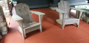 Two unpainted Adirondack chairs facing a us with the homes kitchen seen in the background. These chairs are in an outdoor shelter. The concrete floor is painted grand Canyon red. Humming Bird Exclusives.